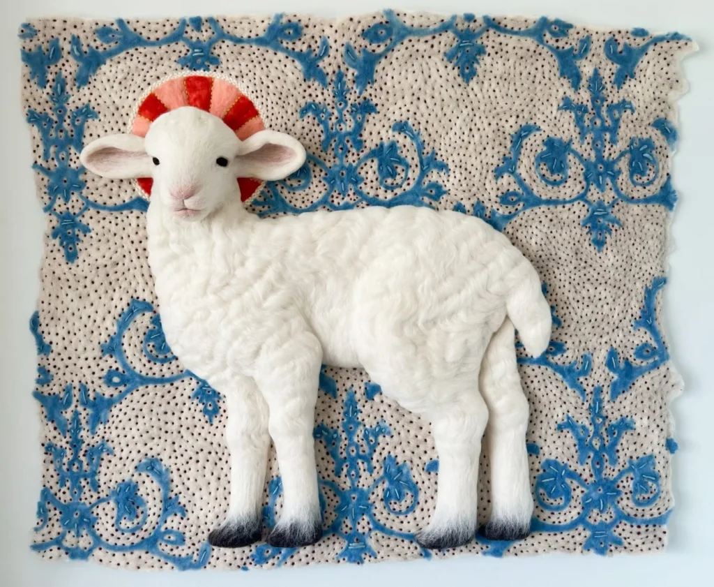 Needle-felted white lamb sculpture displayed in profile against an ornate blue botanical felt tapestry. A circular red-and-pink halo-like motif frames the lamb's head, while scrolling floral patterns decorate the cream-colored textile backdrop. The lamb's textured fleece, soft facial features, and dark hooves create a striking contrast with the intricate handcrafted background, producing a contemporary folk art composition