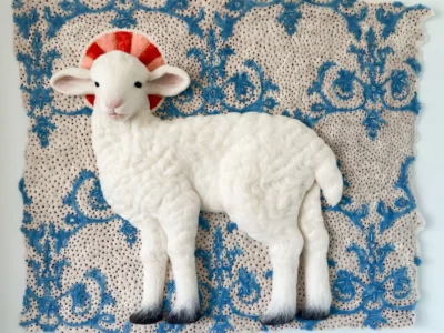 Needle-felted white lamb sculpture displayed in profile against an ornate blue botanical felt tapestry. A circular red-and-pink halo-like motif frames the lamb's head, while scrolling floral patterns decorate the cream-colored textile backdrop. The lamb's textured fleece, soft facial features, and dark hooves create a striking contrast with the intricate handcrafted background, producing a contemporary folk art composition
