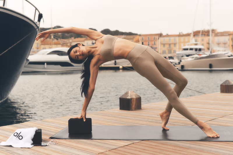 A woman performs a yoga pose on a waterfront dock beside luxury yachts, dressed in a neutral-toned Alo workout set. The serene harbor setting emphasizes balance, flexibility, and mindful movement within a haute wellness lifestyle