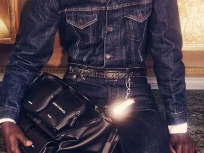 Close-up campaign image from Maison Christian Louboutin Fall/Winter 2026 menswear featuring a dark indigo denim jacket and jeans, branded Christian Louboutin belt, and a structured black leather utility bag with multiple zippered compartment