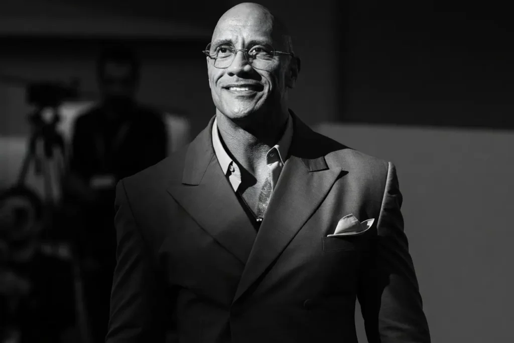 Dwayne Johnson in a tailored gray suit and glasses, smiling confidently — black and white editorial portrait for his 2026 career evolve