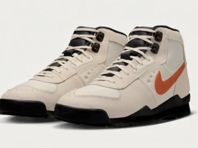Nike hiking-inspired mid-top boots in sail and cream leather with bright orange Swooshes, black rugged outsoles, metal lace hooks, and outdoor-ready heritage style