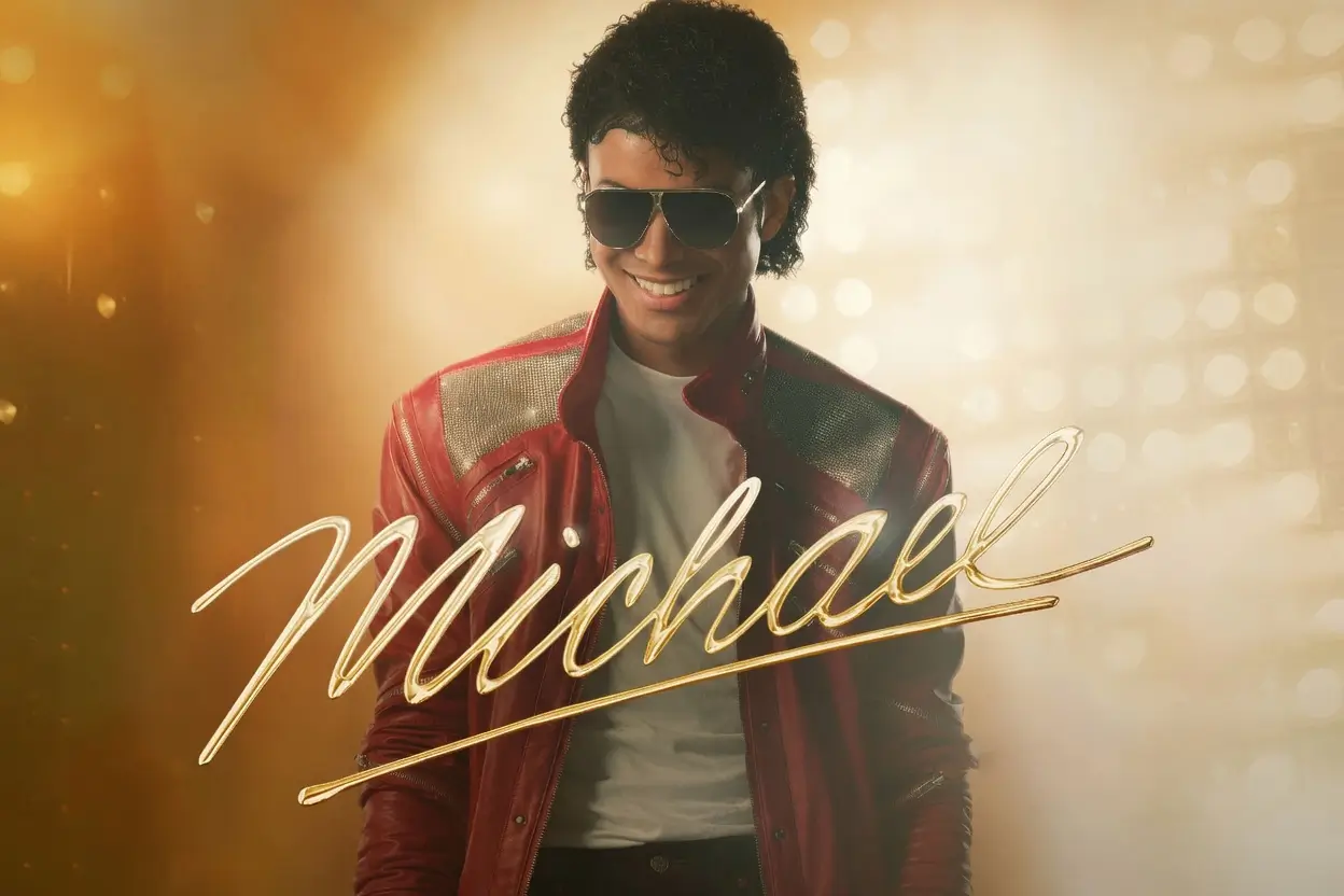 A cinematic promotional poster features a performer portraying the legendary pop icon in a red leather jacket, white T-shirt, and aviator sunglasses. Warm golden lighting and soft bokeh effects create a glamorous concert-like atmosphere, while a large gold cursive signature reading “Michael” stretches across the foreground. The composition evokes the energy, style, and enduring legacy associated with one of music’s most influential entertainer