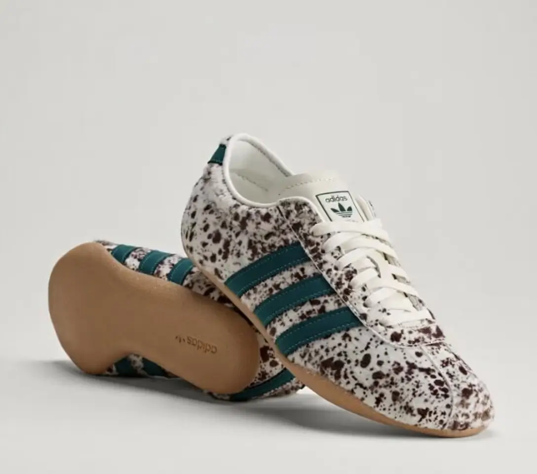 Angled product shot of adidas shoe with a white and brown speckled finish, teal side stripes, and a classic gum outsole. One shoe is positioned upright while the other displays the sole, highlighting the minimalist silhouette and vintage indoor-sport aesthetic