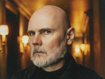 Close-up portrait of a Billy Corgan with a neatly trimmed gray beard and intense expression, photographed in a warmly lit interior corridor. Soft amber wall lamps line the hallway behind him, creating a cinematic glow and shallow depth of field. The subject wears dark clothing, while the blurred background and subtle film grain lend the image a moody, atmospheric quality
