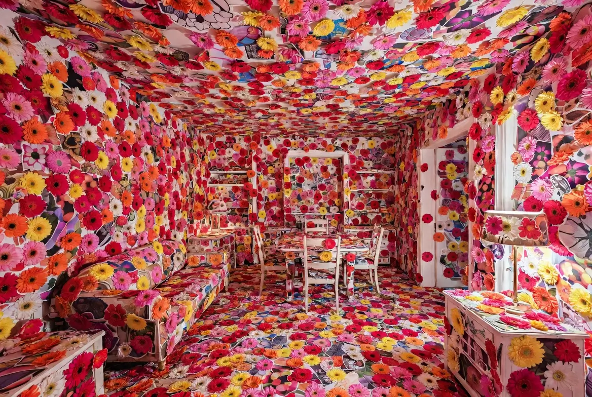 Immersive floral installation room covered floor-to-ceiling with multicolored gerbera flowers, transforming furniture, walls, ceiling, and décor into a vibrant contemporary art environment bursting with tincture and pattern