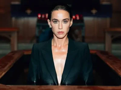 Kate Perry with slicked-back dark hair and a serious expression emerges from a wooden baptismal-style pool, wearing a deep-plunge black tailored blazer. The dimly lit church-like interior creates a dramatic, cin atmosphere