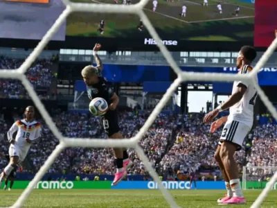 View from behind the goal captures a Paraguayan attacker leaping to head the ball toward goal during a soccer match against Germany. The white goal net frames the action, with German defenders in white kits and pink boots watching the play. A packed stadium, giant video screen, and advertising boards create an energetic international tournament atmosphere