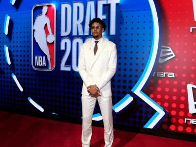 A person stands on the red carpet at the 2026 NBA Draft wearing a white double-breasted suit, white shirt, dark tie, and tinted sunglasses. The colorful NBA Draft backdrop with NBA and New Era branding fills the background