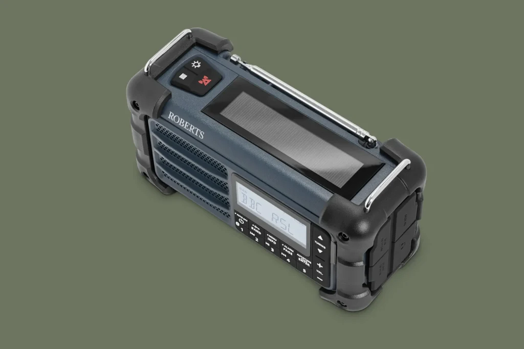 Three-quarter top view of a rugged portable emergency radio in matte navy blue and black. The device features a large solar panel across the top, a telescopic antenna, reinforced rubberized corner bumpers, and an illuminated LCD display with physical control buttons. Designed for outdoor use, the compact radio combines weather-ready durability with modern functionality against a muted olive-green studio background