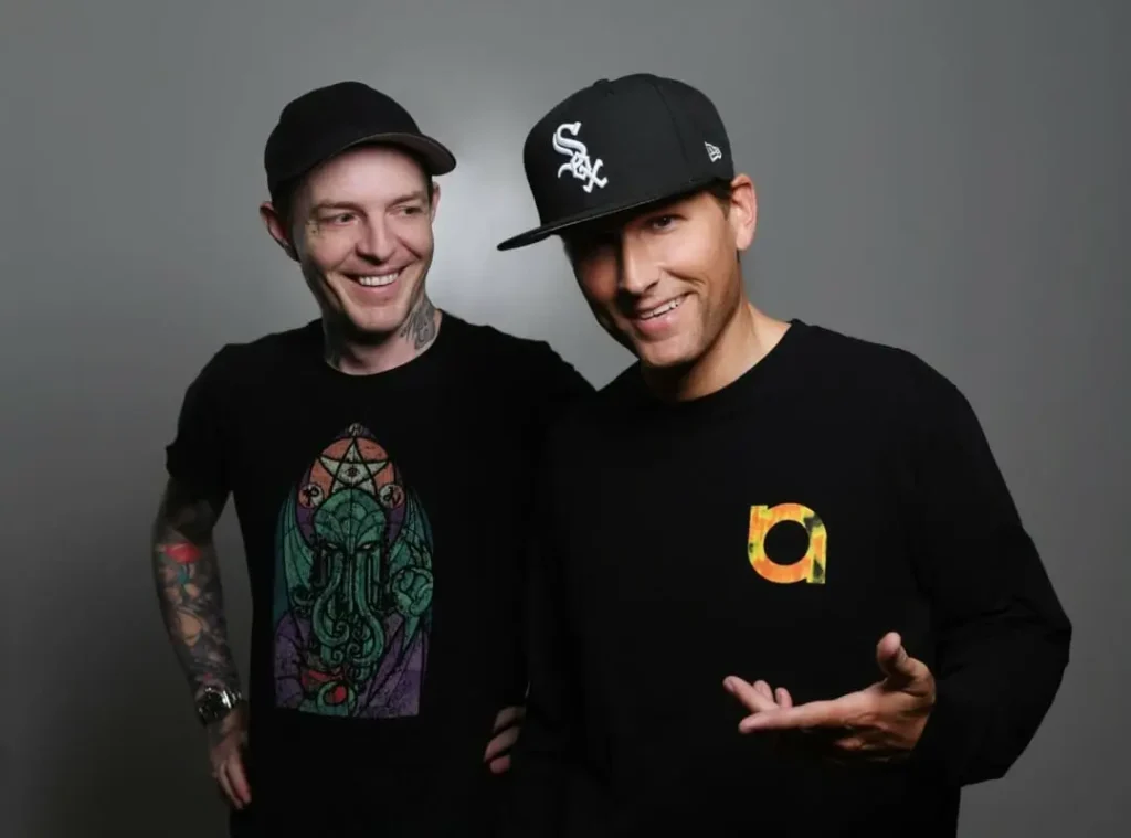 Studio portrait of Kx4 wearing black baseball caps and black graphic shirts, posing against a neutral gray background. One stands slightly behind while the other gestures toward the camera in a relaxed, friendly pose