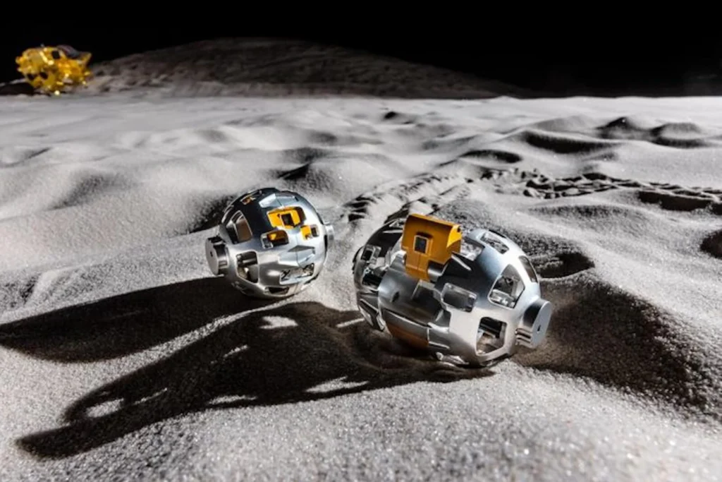 Two spherical autonomous lunar robots traverse the Moon’s dusty surface, casting long shadows across rugged regolith. In the distance, a lunar lander sits near the horizon beneath the stark blackness of space, highlighting a futuristic vision of robotic lunar exploration and off-world mobile