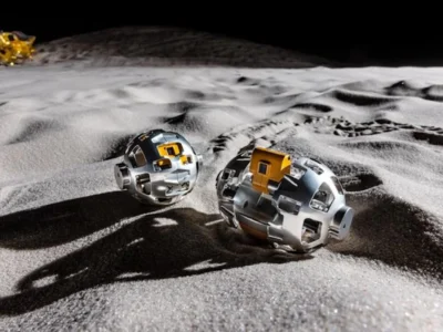 Two spherical autonomous lunar robots traverse the Moon’s dusty surface, casting long shadows across rugged regolith. In the distance, a lunar lander sits near the horizon beneath the stark blackness of space, highlighting a futuristic vision of robotic lunar exploration and off-world mobile
