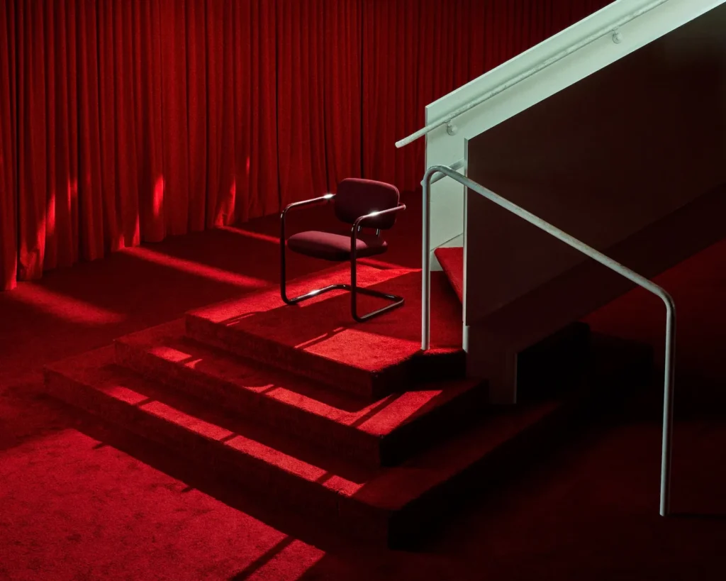 Dramatic red-lit scene featuring a SOM79-style cantilever chair on red-carpeted stairs, referencing Halston’s signature all-red Olympic Tower aesthetic and theatrical glamour