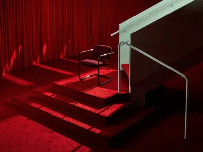 Dramatic red-lit scene featuring a SOM79-style cantilever chair on red-carpeted stairs, referencing Halston’s signature all-red Olympic Tower aesthetic and theatrical glamour