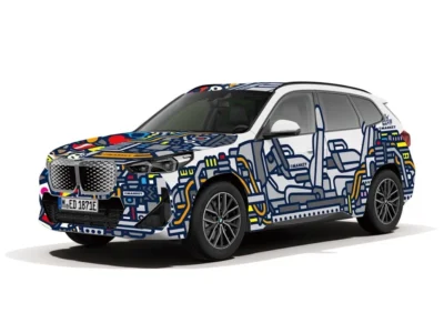 Three-quarter front view of a BMW SUV displayed against a light gray studio background, wrapped in a bold graphic art livery. The vehicle features intricate blue line illustrations across the doors and body panels, accented with yellow, red, pink, and white geometric shapes on the hood, roof, and front fascia. Large alloy wheels, BMW’s illuminated kidney grille, and the playful mural-style design create a striking blend of automotive engineering and contemporary street-art aesthetic
