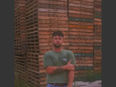 Portrait of Public Works musician Jacob Dietrich standing with arms crossed in front of towering stacks of weathered wooden pallets. The image features a warm, grainy aesthetic with muted earth tones, highlighting the DIY, working-class atmosphere that reflects the raw and heartfelt character of his emo-folk and punk-inspired music project