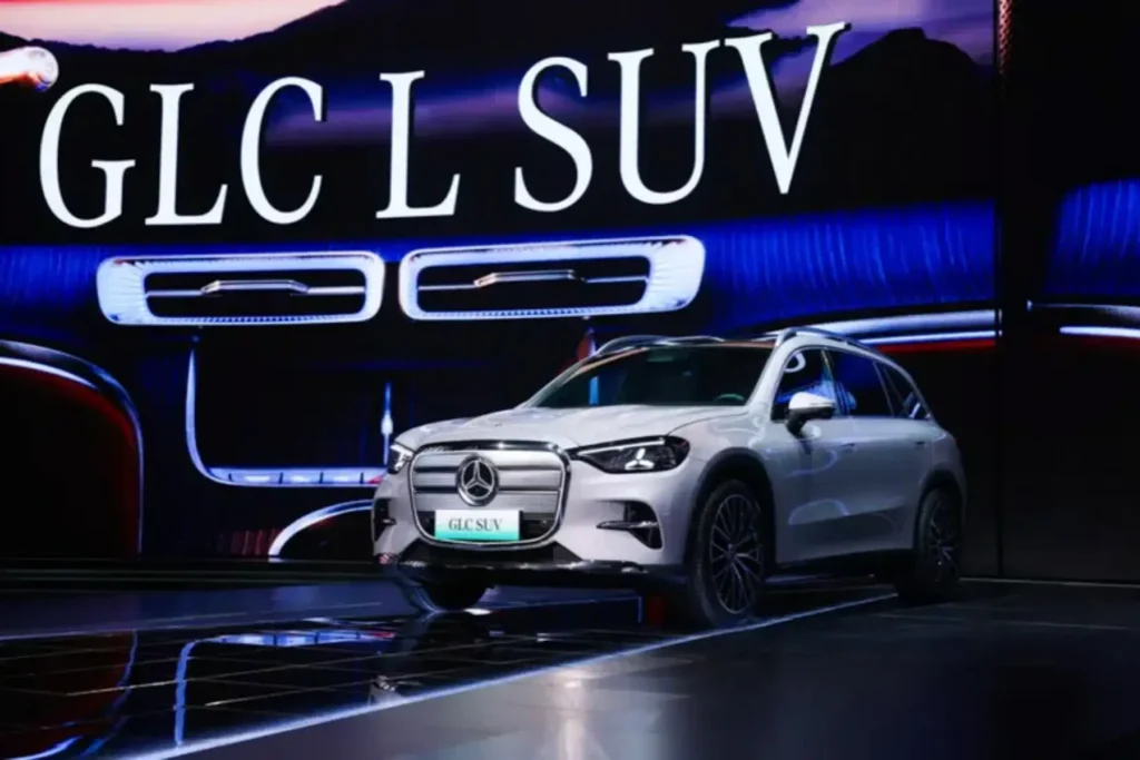 A white Mercedes-Benz GLC electric SUV is displayed on a futuristic stage during its unveiling event. Behind the vehicle, a massive illuminated screen reads “GLC SUV” and highlights the model’s signature dashboard design with blue ambient lighting and rectangular air vents. Reflections across the glossy floor and dramatic lighting emphasize the vehicle’s modern electric styling and premium luxury position