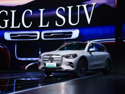 A white Mercedes-Benz GLC electric SUV is displayed on a futuristic stage during its unveiling event. Behind the vehicle, a massive illuminated screen reads “GLC SUV” and highlights the model’s signature dashboard design with blue ambient lighting and rectangular air vents. Reflections across the glossy floor and dramatic lighting emphasize the vehicle’s modern electric styling and premium luxury position