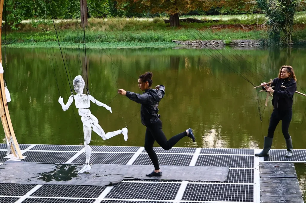 A performer in black athletic wear mirrors the movements of a suspended white marionette sculpture on a floating stage beside a calm lake. Another puppeteer stands nearby controlling the figure with strings, while lush green trees and reflections create a theatrical outdoor set