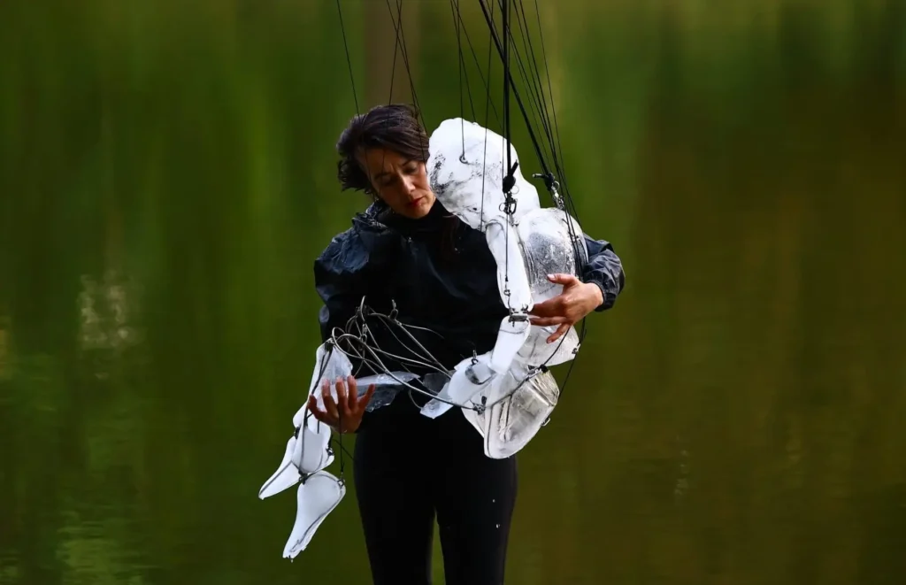 A performer gently cradles a white articulated puppet against their chest in front of a serene green lake. Thin suspension wires frame the sculpture’s limbs and torso, emphasizing the emotional connection between human and puppet during the contemporary show