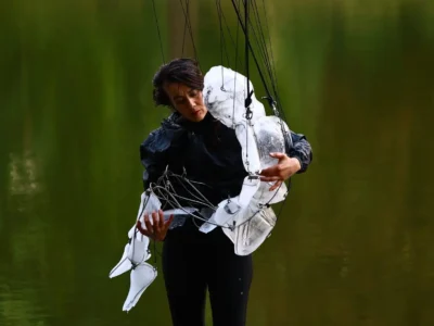 A performer gently cradles a white articulated puppet against their chest in front of a serene green lake. Thin suspension wires frame the sculpture’s limbs and torso, emphasizing the emotional connection between human and puppet during the contemporary show