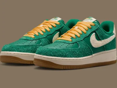 A pair of Nike Air Force 1 “Game Day” shoe is photographed from a three-quarter angle, showcasing the combination of textured suede, smooth leather panels, and vintage-inspired color blocking. The gum sole and cream Swoosh provide a balanced contrast against the vibrant green upper