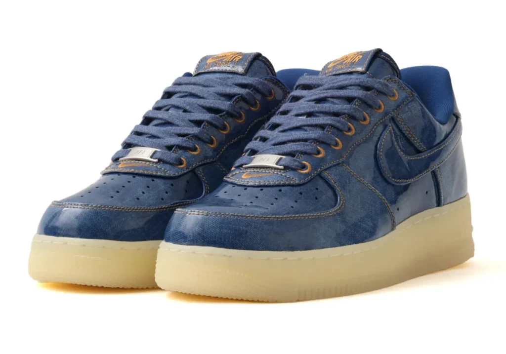 Angled front-quarter view of the Nike Air Force 1 Low Patent Denim, highlighting the glossy blue denim-inspired upper, gold contrast stitching, metal lace dubraes, and vintage-tinted translucent sole unit