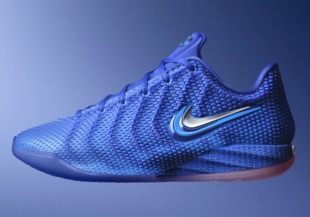 Side profile of a royal blue Nike Caitlin Clark signature basketball shoe against a matching blue gradient background. The low-top silhouette features a textured woven upper, tonal laces, a metallic blue-and-silver Swoosh, and a sculpted performance midsole designed for speed, stability, and on-court respond