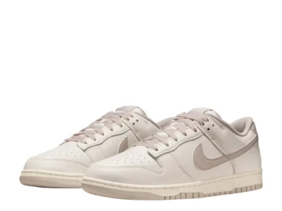 A pair of Nike Dunk Low sneakers is displayed at a three-quarter angle, emphasizing the perforated toe boxes, tonal branding, and minimalist neutral color scheme