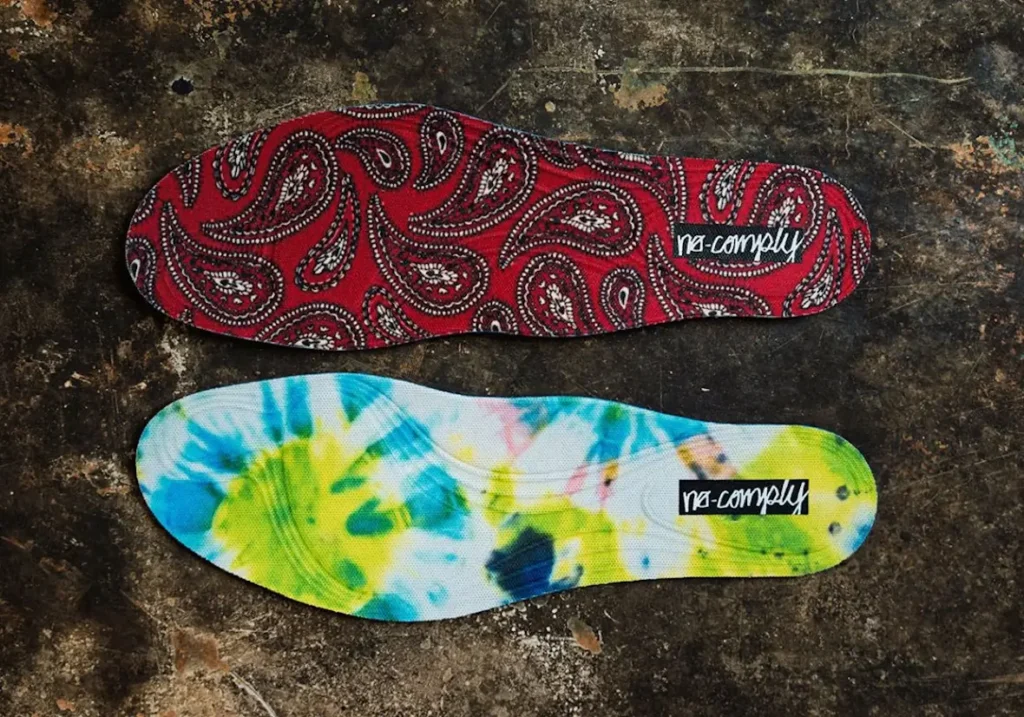 Two removable insoles are presented side by side, one featuring a red paisley print and the other a colorful tie-dye pattern, both finished with No-Comply brand