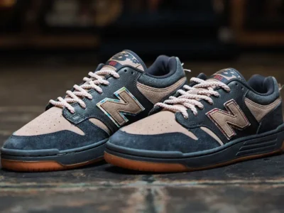 A pair of No-Comply x New Balance Numeric 480 sneakers is displayed on a polished floor, showcasing black suede mudguards, tan suede uppers, mismatched logo detailing, and gum rubber outsole