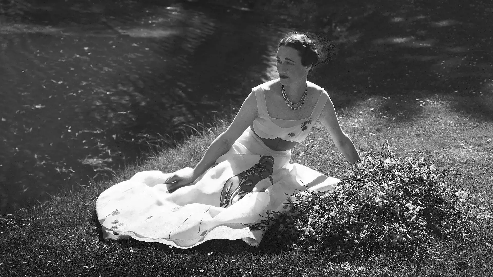 Black-and-white portrait of a woman seated beside a pond in a lobster-print dress, surrounded by grass and blooming branches in a serene outdoor set