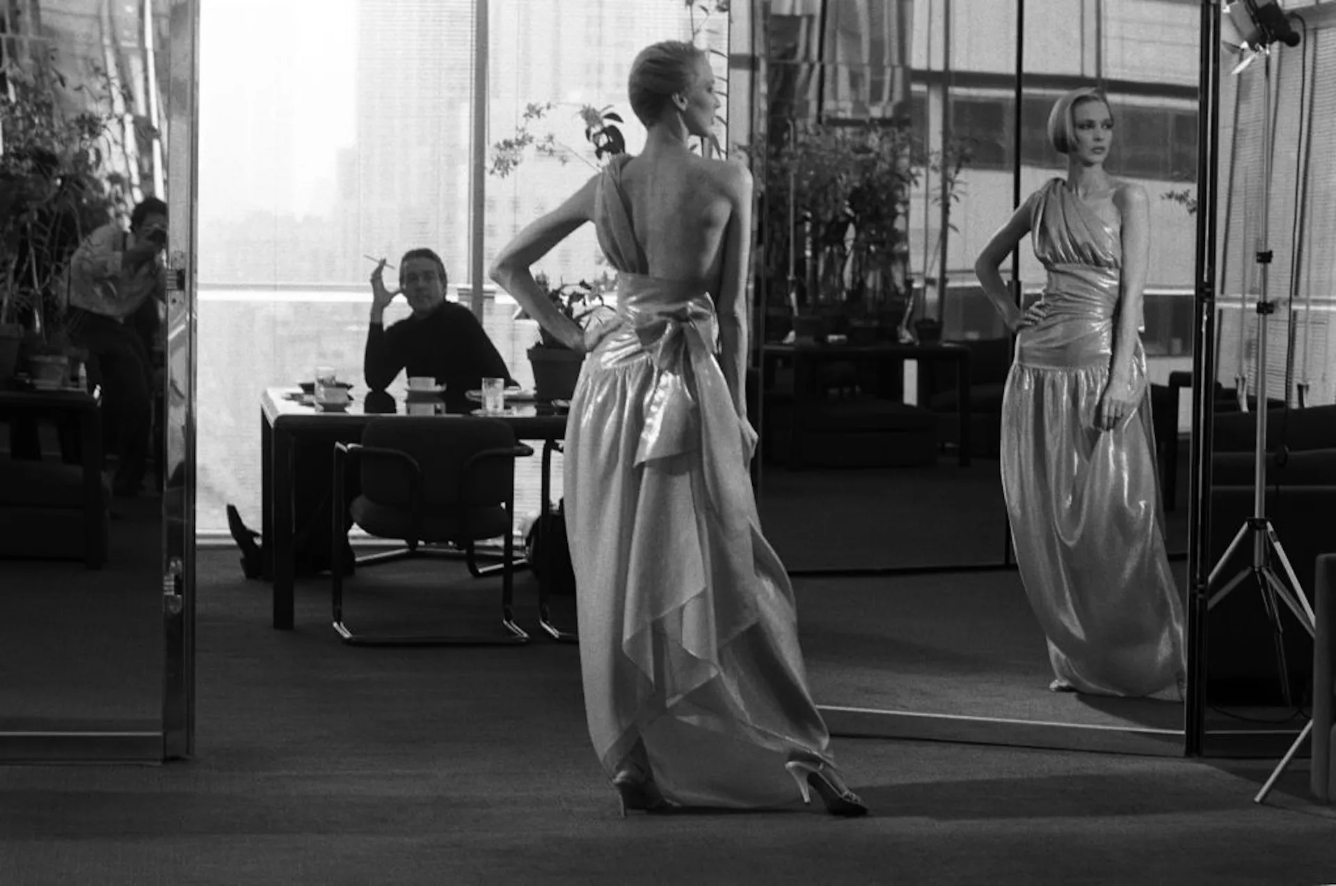 Black-and-white fashion editorial featuring a model in a metallic, backless gown posing before a mirrored wall in a modern office. Reflections, soft lighting, and a behind-the-scenes studio setting create a sophisticated luxury-fashion atmosphere
