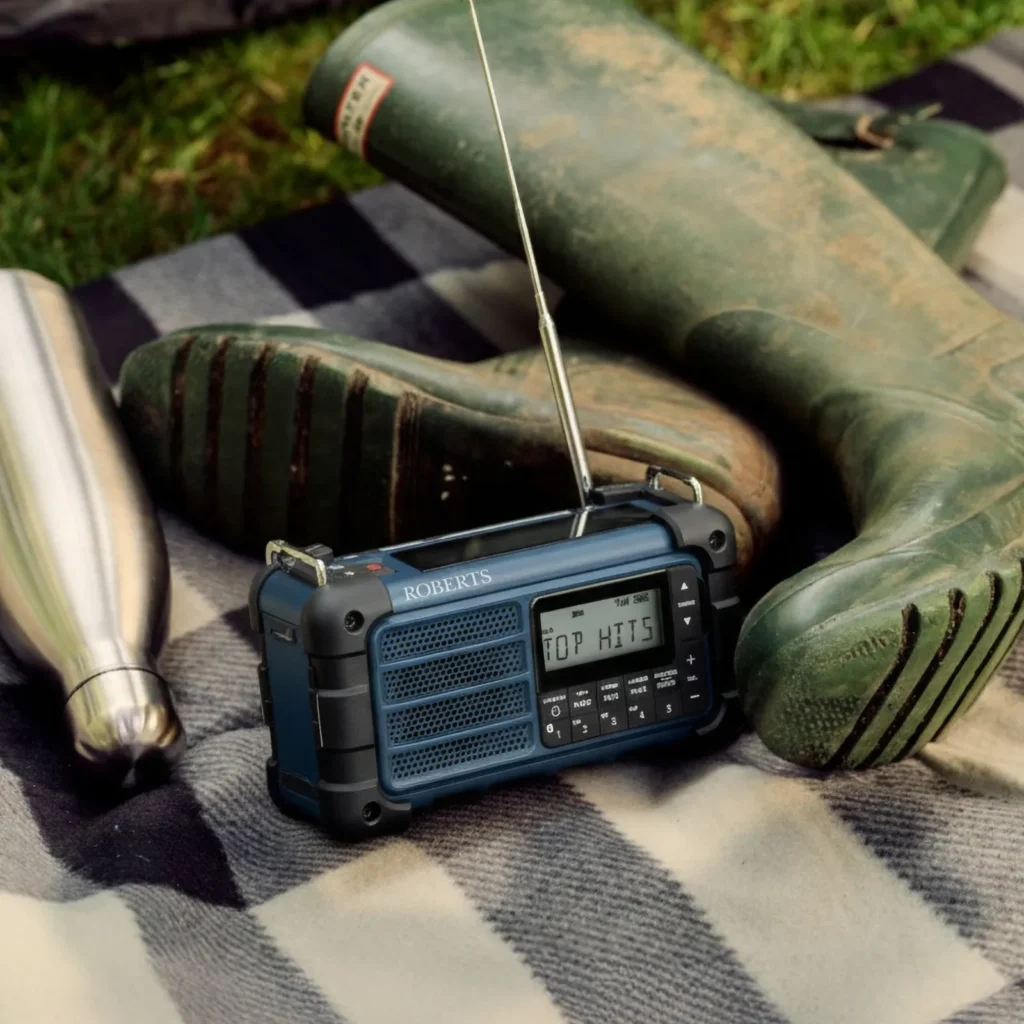 Lifestyle image of the rugged portable radio resting on a black-and-white picnic blanket outdoors beside worn green Wellington boots and a stainless steel water bottle. The extended antenna and illuminated display reinforce its camping and emergency-use credentials, while natural lighting adds an authentic outdoor adventure atmosphere
