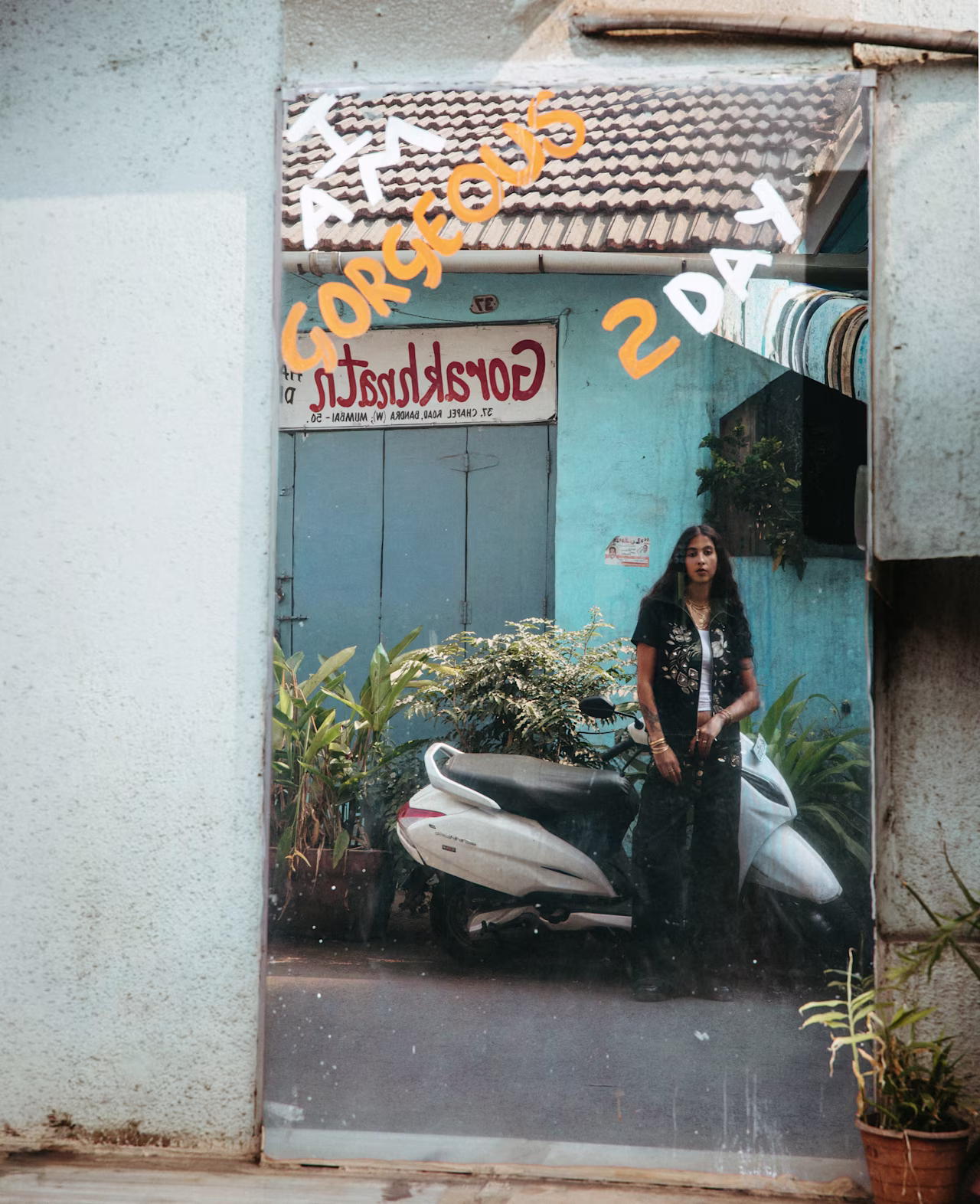 A reflective street-style image captured through a weathered mirror. The model stands beside a white scooter in front of a blue storefront, while handwritten text on the glass and subtle imperfections create a layered, cinematic composition