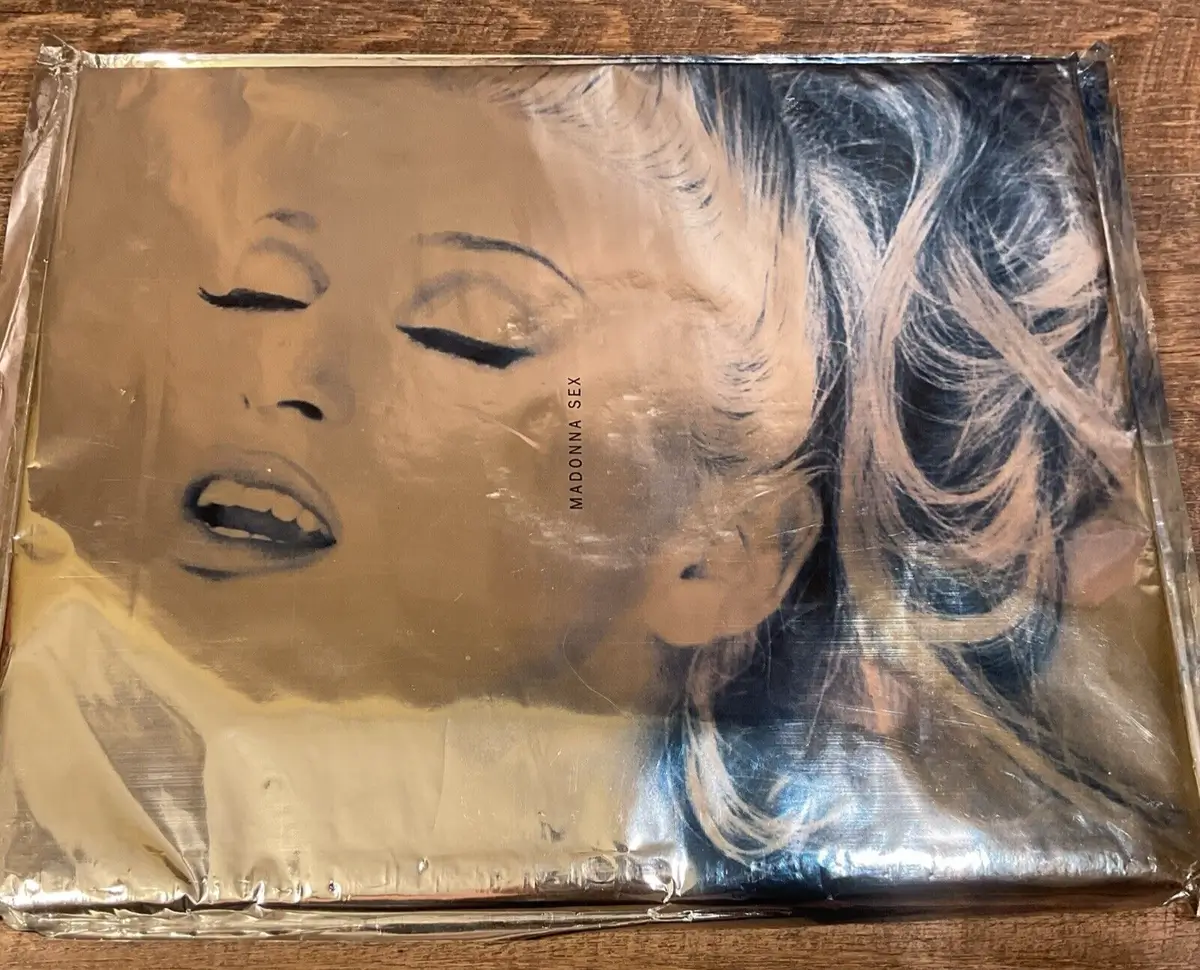 A sealed copy of Madonna’s controversial 1992 book Sex rests on a wooden surface, wrapped in its original metallic silver Mylar packaging. The cover features a close-up black-and-white portrait of Madonna with closed eyes and parted lips, highlighting one of the most provocative and culturally influential publications of the early 1990s