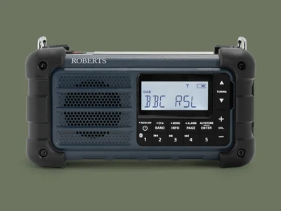 Front-facing studio shot of the compact emergency radio featuring a perforated speaker grille, monochrome LCD display, dedicated tuning controls, preset buttons, and durable corner protection. The navy-blue and black color scheme gives the radio a functional, industrial appearance suited for emergency preparedness and outdoor recreation