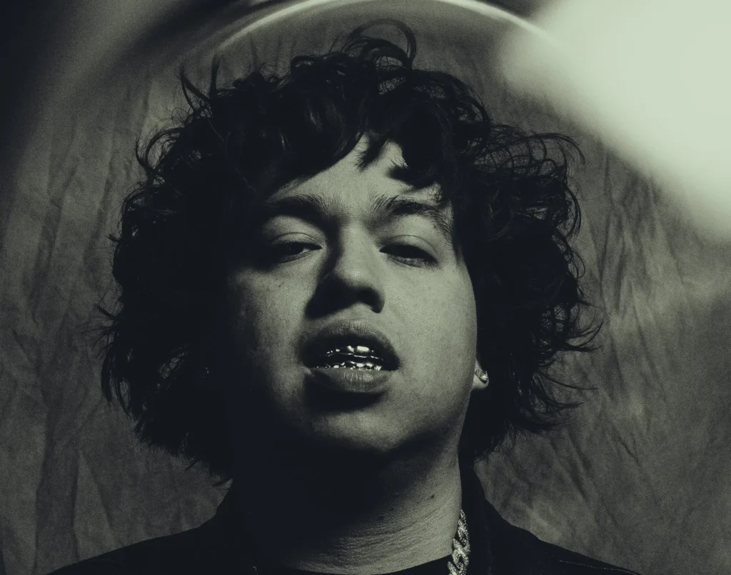 Moody black-and-white portrait featuring 2slimey with tousled curls, metallic grills, and layered jewelry, captured from a low angle with dramatic lighting and a grainy editorial aesthetic