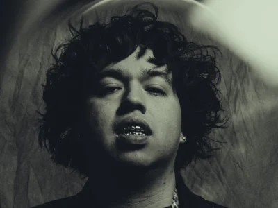 Moody black-and-white portrait featuring 2slimey with tousled curls, metallic grills, and layered jewelry, captured from a low angle with dramatic lighting and a grainy editorial aesthetic