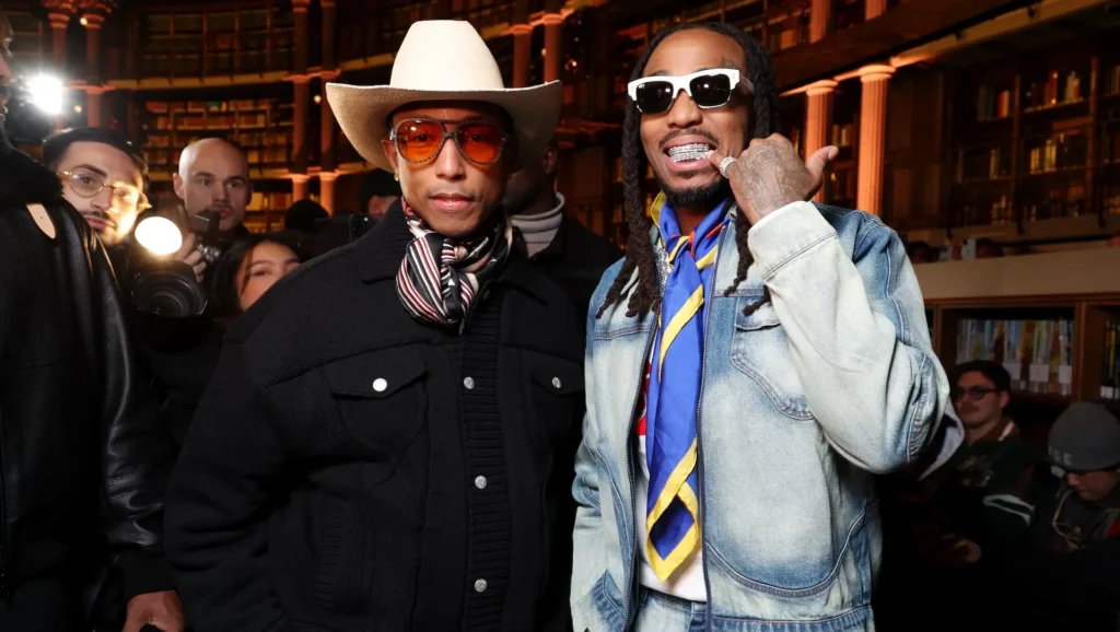 Two stylish attendees pose inside a grand library-like venue with towering bookshelves and warm amber lighting. One wears a cream cowboy hat, orange-tinted glasses, a striped scarf, and a black textured jacket, while the other sports white sunglasses, a faded denim jacket, a colorful silk scarf, and layered jewelry. Photographers and guests surround them, creating a lively fashion-week atmosphere