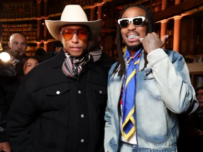 Two stylish attendees pose inside a grand library-like venue with towering bookshelves and warm amber lighting. One wears a cream cowboy hat, orange-tinted glasses, a striped scarf, and a black textured jacket, while the other sports white sunglasses, a faded denim jacket, a colorful silk scarf, and layered jewelry. Photographers and guests surround them, creating a lively fashion-week atmosphere