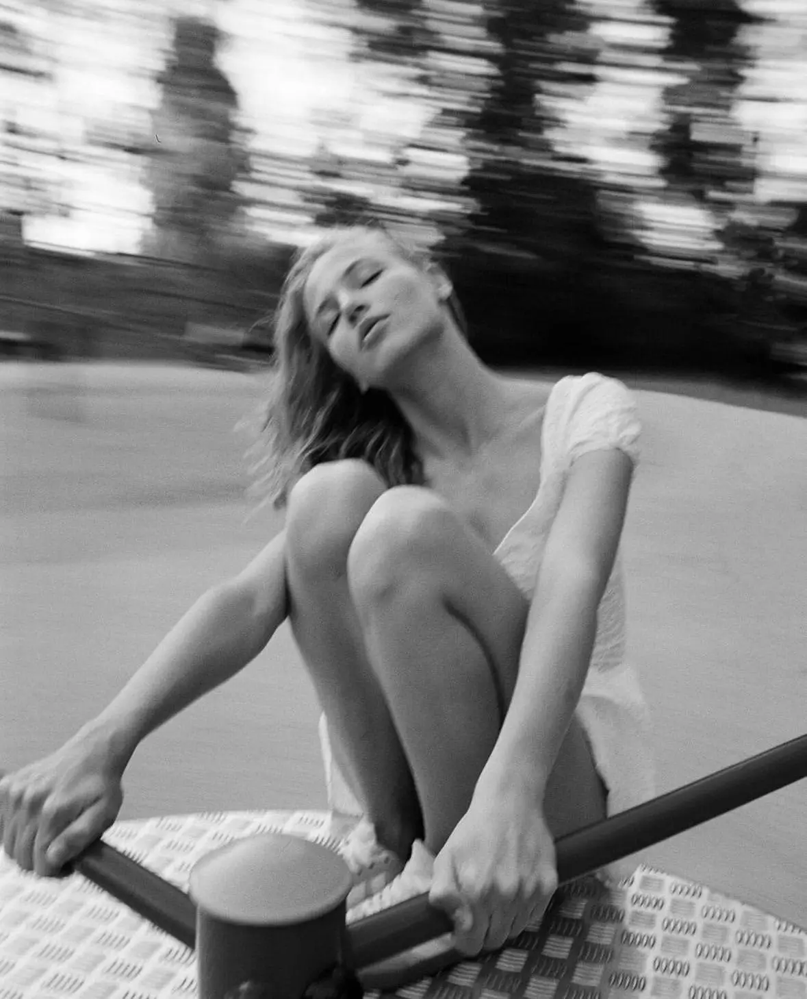 Black-and-white motion-blur photograph of a woman seated on a playground roundabout, eyes closed and head tilted back, creating a dreamy, nostalgic sense of freedom and move