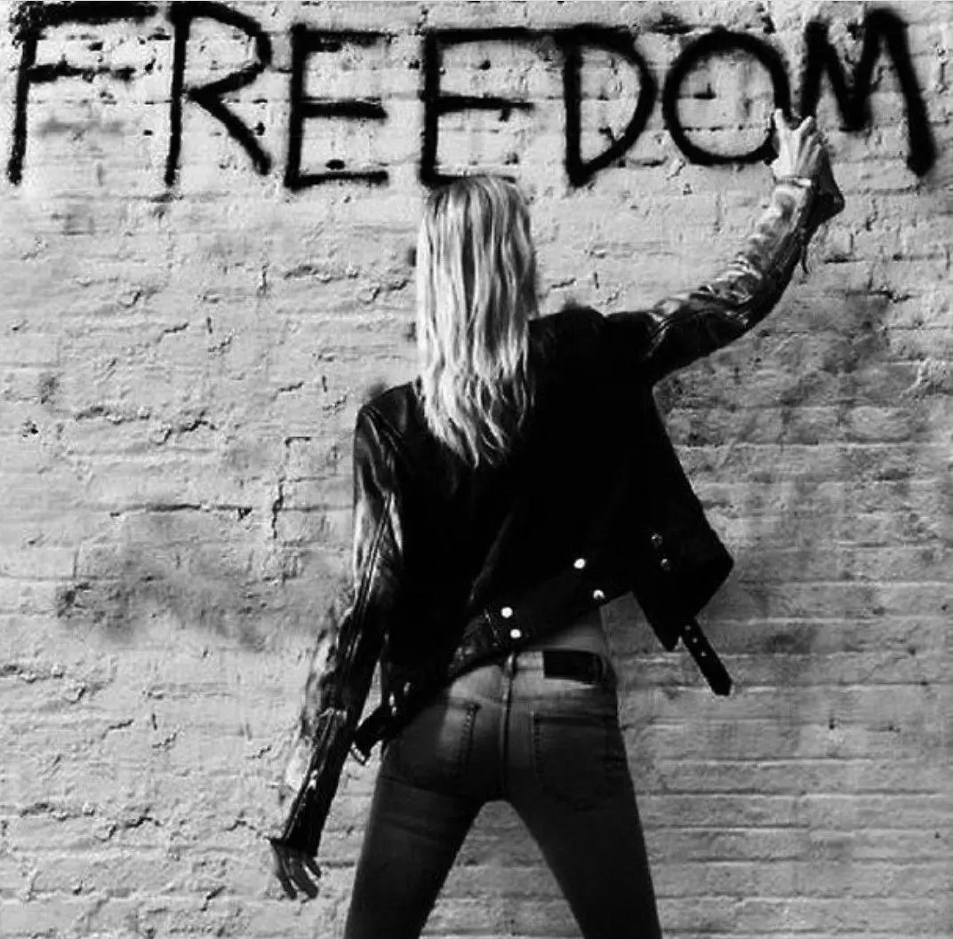 Black-and-white photograph of a woman in a leather jacket spray-painting the word “FREEDOM” across a weathered brick wall in an urban set