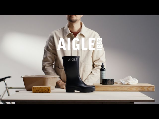 A studio lifestyle scene showcases a black AIGLE rubber rain boot standing on a white table beside cleaning supplies, including a basin, sponge, cloth, and bottle. A person wearing a light beige work jacket stands behind the display, while the large “AIGLE 1853” logo appears prominently in the background. The minimalist setting emphasizes the boot’s durable construction, tall shaft, and practical outdoor-inspired design
