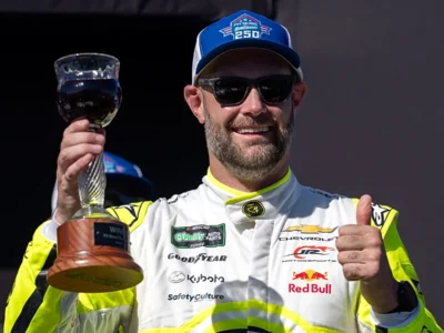 Race-winning driver celebrates on the podium wearing a white and neon yellow Chevrolet racing suit, blue event cap, and sunglasses while holding a trophy aloft and giving a thumbs-up, with sponsor logos including Red Bull, Goodyear, O'Reilly Auto Parts, and Kubota view on the suit
