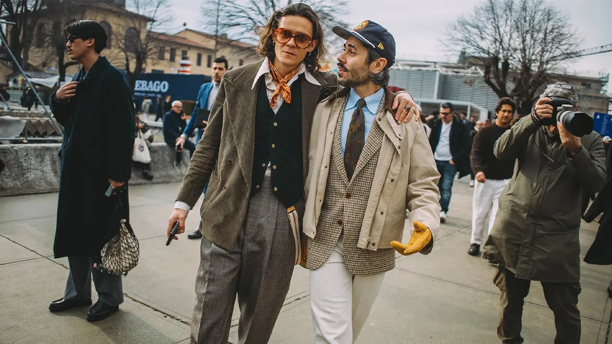 Street style attendees arrive at Milan Fashion Week Menswear Spring/Summer 2027 wearing tailored layers, vintage-inspired suiting, scarves, ties, and oversized outerwear while photographers capture the scene outside a show venue