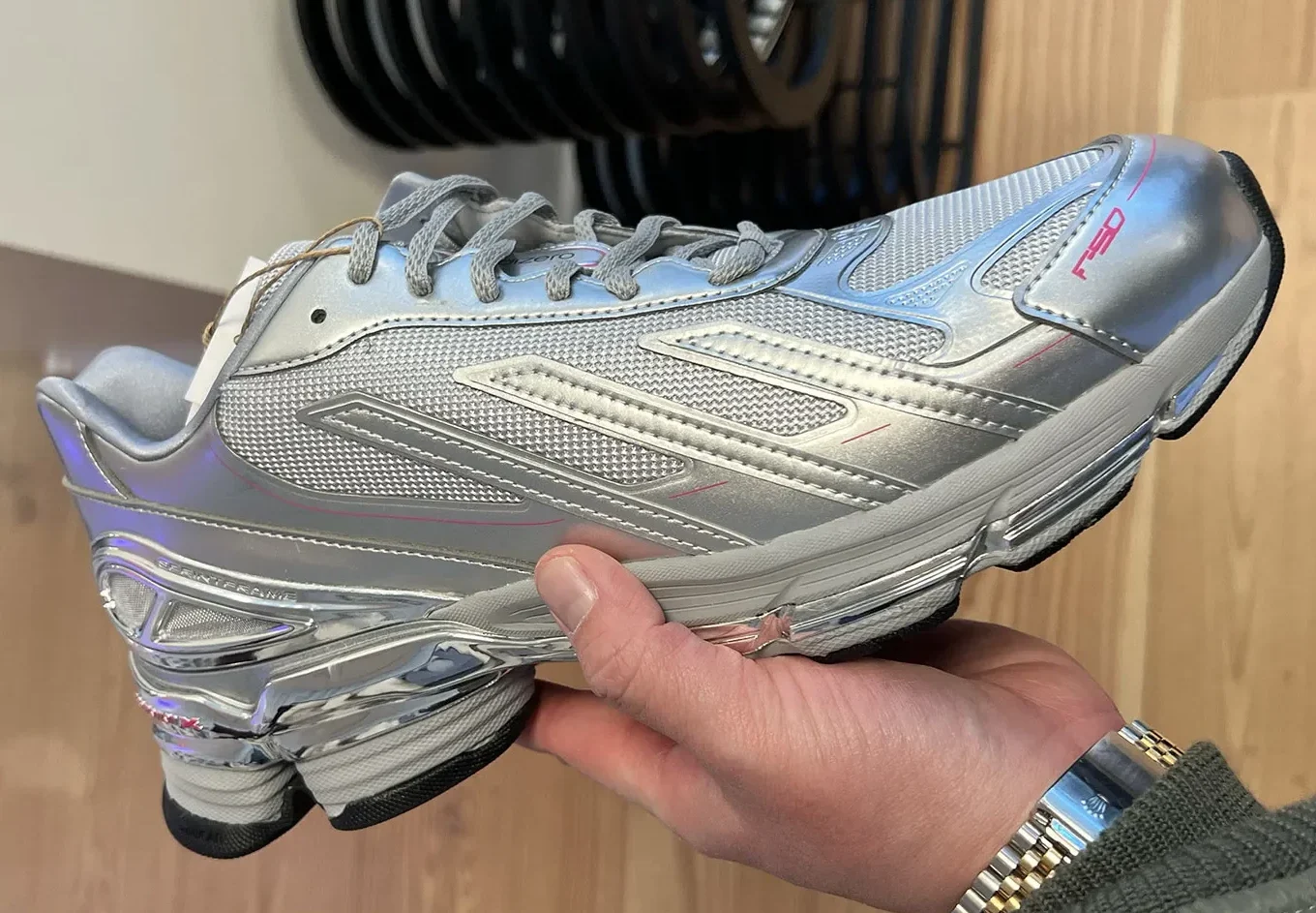 Hand-held side profile of the Sp5der x adidas F50 Formotion “Silver Metallic” sneaker inside a retail setting. The shoe features a metallic silver upper with breathable mesh panels, reflective chrome Formotion heel tooling, grey laces, subtle pink graphic accents, and bright pink F50 branding on the toe. A hand wearing a silver-and-gold wristwatch supports the sneaker, while weight plates and wood-paneled walls appear softly blurred in the background