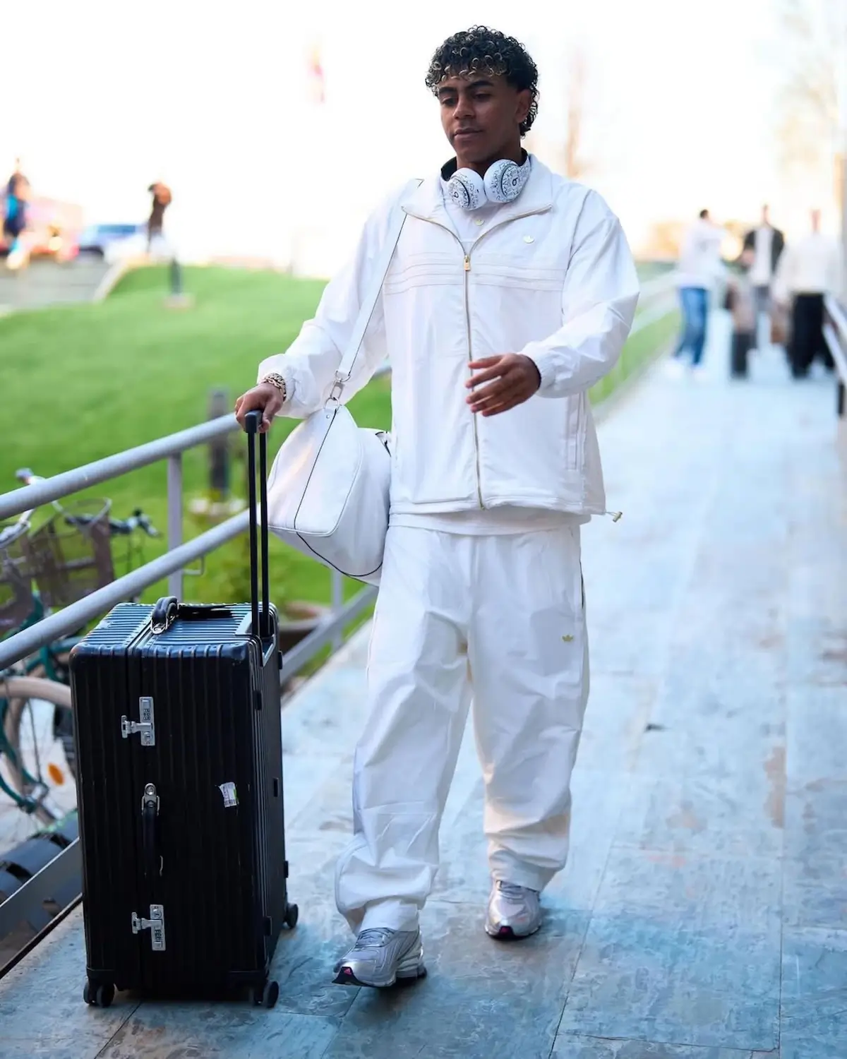 Lifestyle image of a person walking outdoors in an all-white adidas tracksuit while rolling a black hard-shell suitcase. The look is completed with the silver Sp5der x adidas F50 Formotion sneakers and matching white over-ear headphone