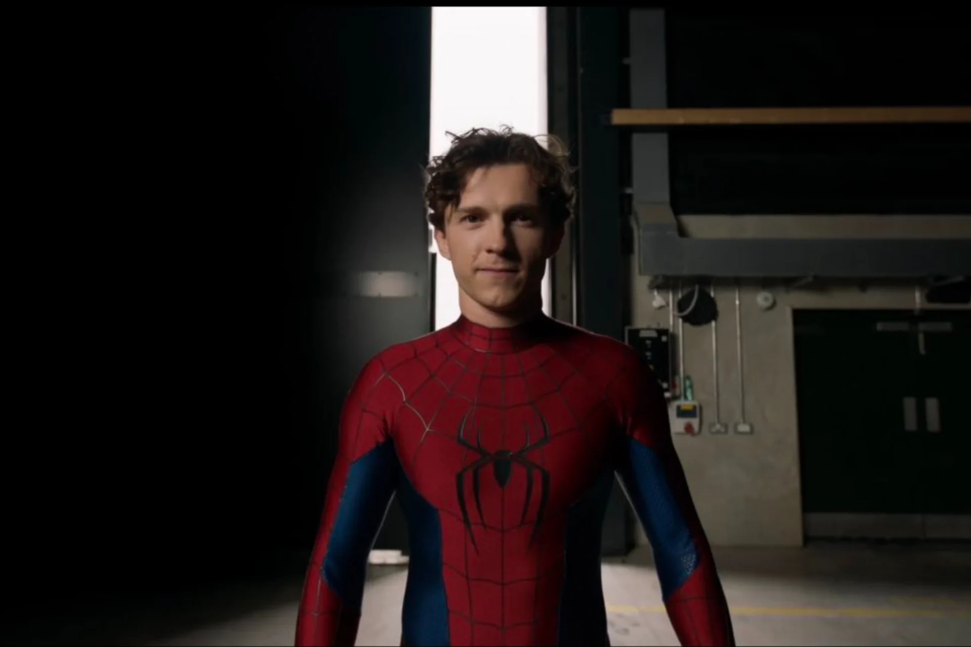 Peter Parker stands in a studio-like setting wearing Spider-Man’s classic red-and-blue suit, with the mask removed to reveal his youthful face and curly hair. A bright vertical light source behind him creates a dramatic silhouette, while the suit’s black spider emblem and web patterning are clearly visible. The scene highlights a more mature and confident version of the hero, emphasizing the return to a traditional comic-inspired costume design